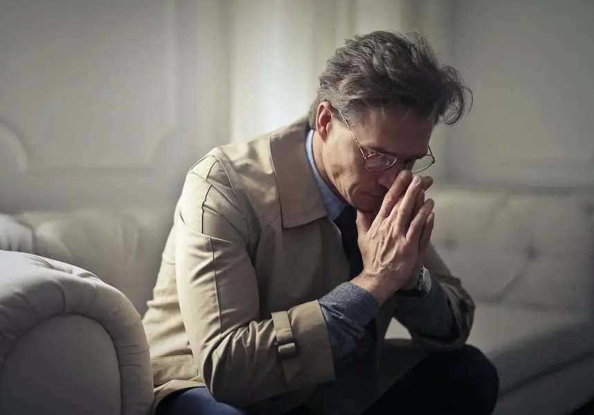 Man sitting on sofa with hands clasped, showing signs of depression or anxiety.