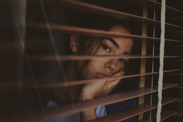 woman looking through window blinds with er head in er hands