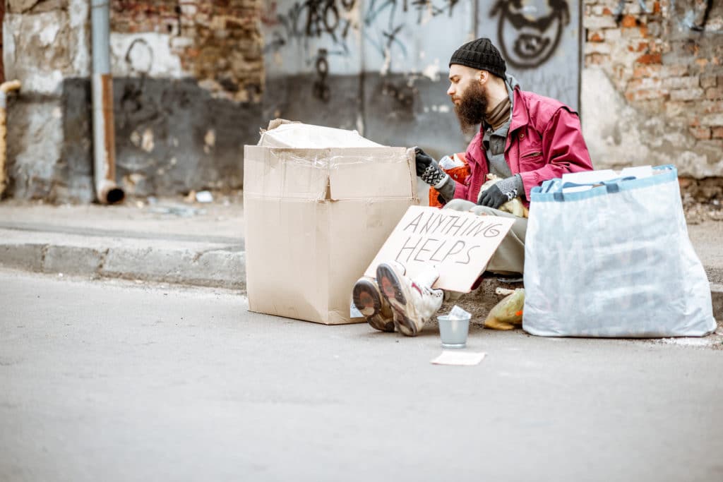 Homeless depressed beggar sitting with bags and cardboards on the street on the ragged wall background. Concept of social poverty