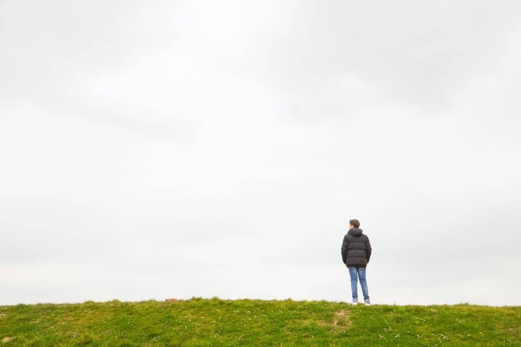 A single person standing on the horizon above a grass field against a large grey clouded sky depicting loneliness, thinking, pondering, grieve, being alone, sadness
