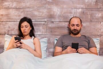 Caucasian young couple under the blanket wearing pajamas using smartphone.