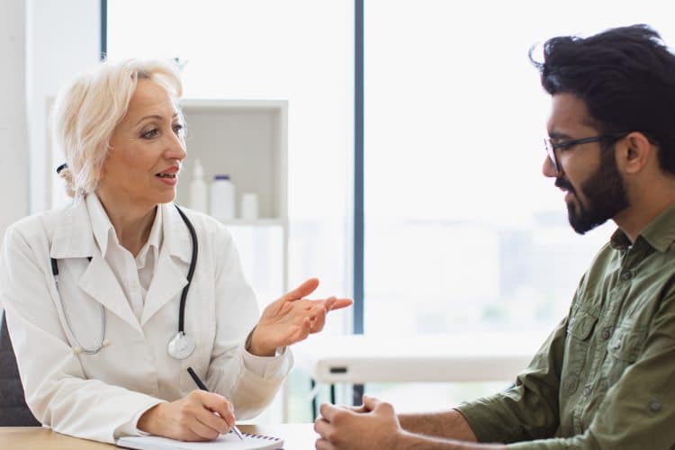 Mature elder physician giving recommendations and using notebook for writing prescription. Bearded gentleman describing symptoms of illness to senior female doctor and gesturing hands.