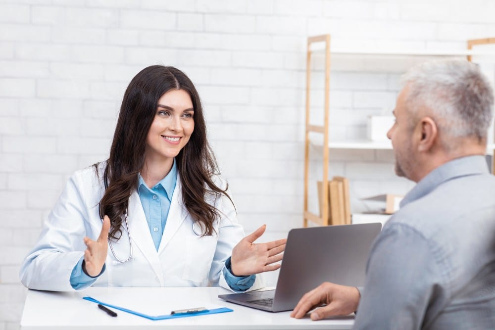 Patient consultation, examination in clinic and medical specialist. Cheerful millennial female doctor in white coat talking with adult man, sitting at table with laptop in office interior,