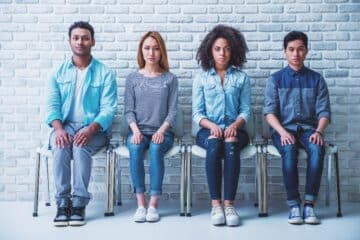 Young people of different nationalities are looking at camera while sitting with hands on knees in a row
