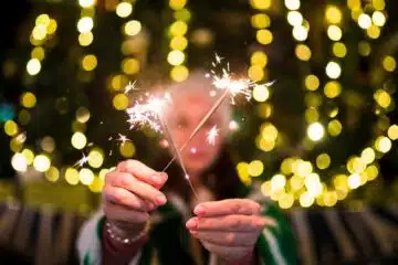 Person holding sparklers with festive lights in background.