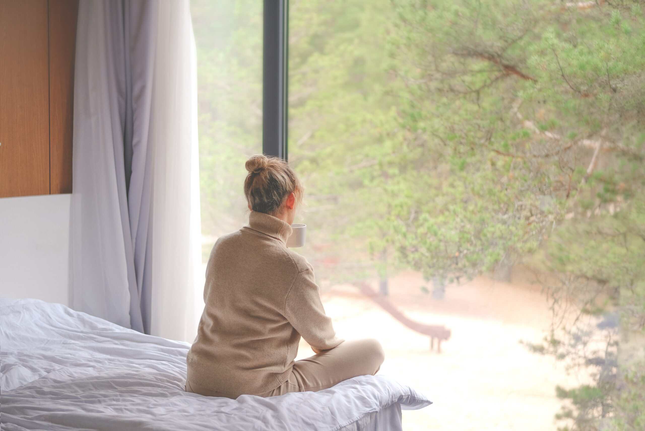 A woman sitting on a bed while looking out the window and drinking coffee for the topic how long is residential treatment for mental health.